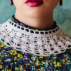 Gros plan d'une femme portant un rouge à lèvres rouge vif, des boucles d'oreilles et un chemisier floral à col haut avec un col au crochet blanc détaillé avec explication sur un fond sarcelle.
