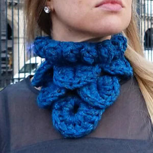 Une femme porte une grosse écharpe bleue crochetée à la main avec des détails en spirale - un parfait col au crochet avec explication. Elle est vêtue d'un haut sombre et se trouve à l'extérieur.