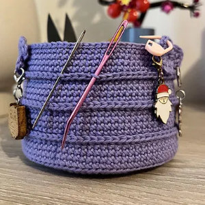 Un panier violet au crochet avec une breloque de Père Noël, un mètre ruban, des ciseaux et des aiguilles est présenté sur une surface en bois - une pièce adorable inspirée par une explication gratuite de panier au crochet.