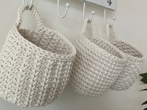 Trois paniers crochetés à la main, de couleur crème et munis de poignées, sont suspendus à des crochets blancs contre un mur clair. Le motif épais et texturé met en valeur leur conception douce et robuste, idéale pour ceux qui recherchent une explication gratuite de panier au crochet.