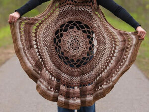 Une personne tend un grand mandala circulaire brun au crochet avec des motifs complexes, présenté sur un fond extérieur avec de la verdure et un chemin pavé.