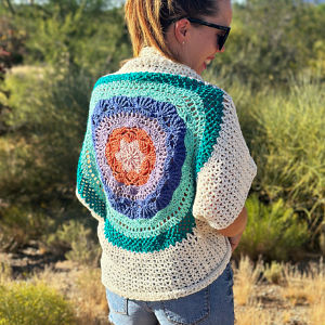 Une femme portant des lunettes de soleil et un pull-over crocheté avec un motif de mandala coloré dans le dos se tient à l'extérieur, dans une zone herbeuse et ensoleillée.