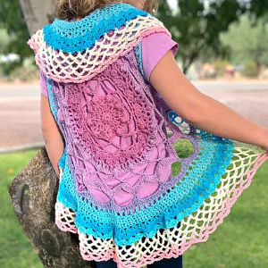 Une personne portant un gilet crocheté coloré avec des motifs complexes dans les tons rose, violet, bleu et blanc, debout à l'extérieur, un bras tendu, montrant le motif circulaire du gilet.
