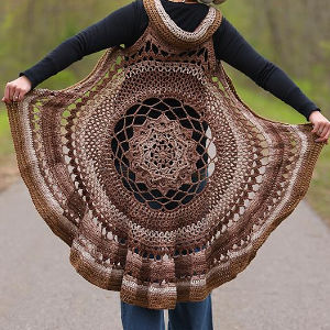 Personne debout en plein air, tendant les côtés d'un grand gilet circulaire en crochet marron avec un motif mandala complexe au centre, créant un effet de cape. L'arrière-plan est un décor naturel flou.