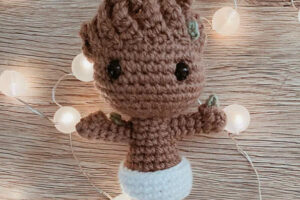 Une poupée au crochet Groot faite à la main se tient sur une surface en bois, entourée de guirlandes lumineuses blanches. Avec ses grands yeux noirs et ses délicats détails verts sur la tête, ce bébé Groot au crochet est à la fois charmant et unique.