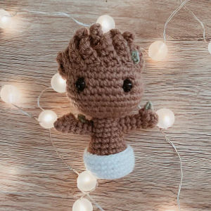 Une figurine crochetée ressemblant à un personnage arborescent avec de grands yeux noirs est posée dans un pot blanc, entourée d'une guirlande de guirlandes lumineuses sur une surface en bois.