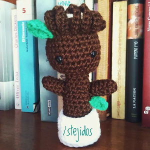 Une poupée brune crochetée avec des feuilles vertes et des yeux noirs se tient devant des livres. La base blanche de la poupée porte l'inscription tejidos en lettres vertes.