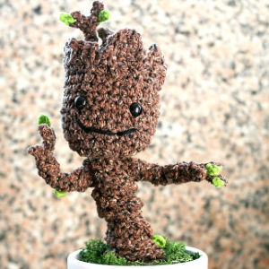 Une figurine crochetée ressemblant à Groot des Gardiens de la Galaxie, avec un visage souriant, un corps texturé marron et de petites feuilles vertes, se tient dans un pot blanc sur un fond flou.