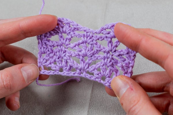 Deux mains tiennent un petit morceau rectangulaire de dentelle au crochet lavande au motif ajouré et dentelé, présenté sur un fond de tissu gris clair.