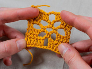 Deux mains tiennent un petit échantillon de crochet jaune avec un motif ouvert et dentelé rappelant la dentelle au crochet, sur un fond de tissu neutre.