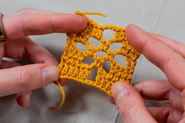 Deux mains tiennent un petit morceau carré de crochet jaune avec un motif ouvert et dentelle rappelant la dentelle au crochet, montrant des rangées de points et des espaces décoratifs. L'arrière-plan est une surface de tissu neutre.