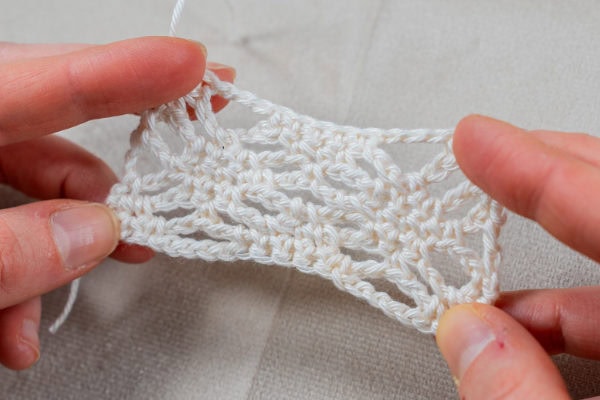 Deux mains tenant un petit morceau de dentelle au crochet blanc, au motif ouvert et aéré, présenté sur une surface texturée de couleur claire.