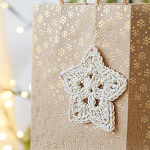 Un sac cadeau marron avec des motifs de flocons de neige dorés est décoré d'une étoile blanche au crochet, réalisée à la main à l'aide d'un modèle d'étoile au crochet gratuit. Des lumières douces et chaudes sont floues en arrière-plan.