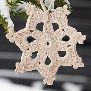 Un flocon de neige beige au crochet - fabriqué à partir d'un modèle d'étoile au crochet gratuit - est suspendu à une branche de conifère saupoudrée de neige sur un fond sombre et flou.