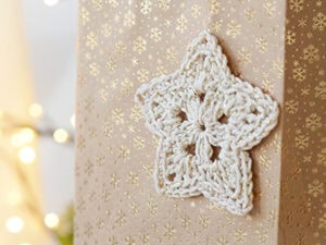 Une étoile blanche au crochet, réalisée à partir d'un modèle d'étoile au crochet gratuit, est attachée à une boîte cadeau dorée décorée de flocons de neige brillants. Des lumières douces et floues brillent en arrière-plan.