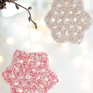 Deux ornements crochetés en forme d'étoile - l'un rose et blanc, l'autre crème - sont suspendus à une branche sur un fond flou et légèrement éclairé, idéal pour mettre en valeur tout modèle d'étoile au crochet gratuit.