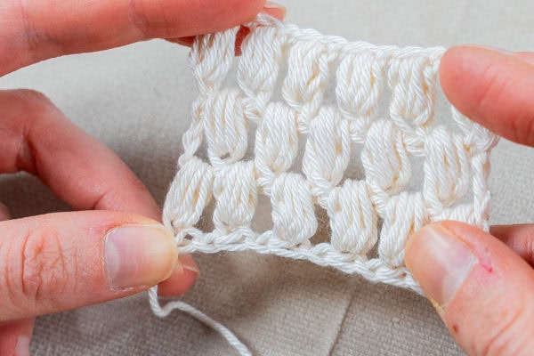 Gros plan de deux mains tenant un petit morceau de tissu blanc au crochet avec des points texturés et verticaux dans le style point ananas au crochet, sur un fond clair.