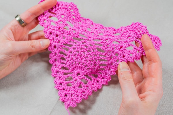 Deux mains tiennent une pièce de dentelle triangulaire au crochet, d'un rose éclatant, réalisée selon la technique du point ananas au crochet, avec des motifs complexes, sur un fond gris clair.