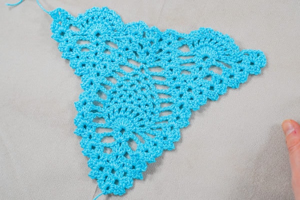 Une pièce de dentelle triangulaire bleu ciel, crochetée à la main, présentant des motifs complexes de points ananas au crochet et de festons, repose à plat sur une surface de tissu léger. Les doigts d'une personne sont visibles dans le coin inférieur droit.