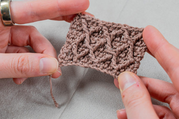 Une personne tient un petit échantillon crocheté en fil marron clair, présentant un motif au crochet texturé et complexe en forme de diamant. Le gros plan met en évidence les mains de la personne et les magnifiques détails au crochet.