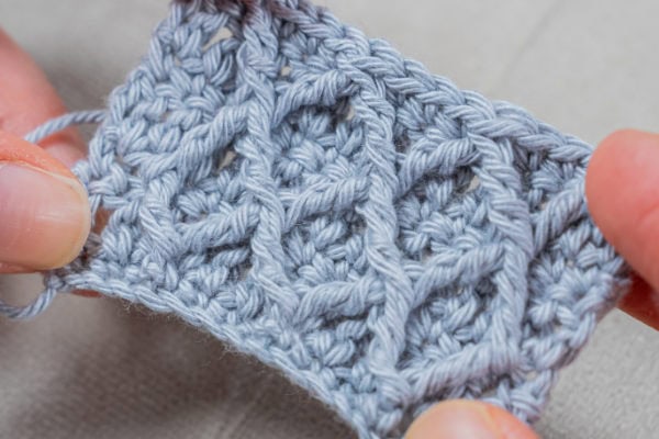 Gros plan de deux mains tenant un petit morceau de tissu crocheté gris clair, mettant en valeur le point diamant au crochet, avec son motif texturé et dentelé composé de lignes diagonales et d'espaces ouverts.