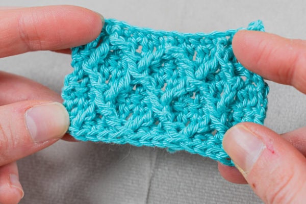 Gros plan de deux mains tenant un petit morceau de tissu au crochet bleu vif, montrant le motif texturé du point diamant au crochet sur un fond neutre.