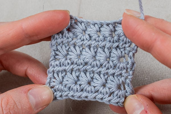 Gros plan de deux mains tenant un petit morceau rectangulaire de tissu crocheté gris clair présentant un motif texturé de point étoile au crochet.