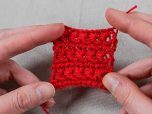 Deux mains tiennent un petit morceau carré de tissu crocheté rouge vif sur un fond gris clair. La pièce présente des points réguliers et nets dans un motif texturé utilisant la technique du point étoile au crochet.
