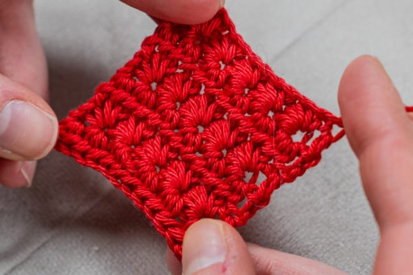 Gros plan des mains tenant un petit échantillon de crochet carré rouge présentant un motif étoile au crochet texturé et en relief. L'arrière-plan est un tissu de couleur claire.