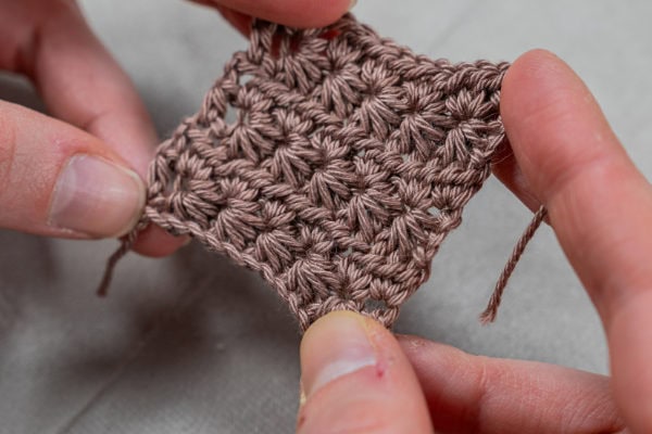 Gros plan des mains tenant un petit échantillon marron crocheté avec le motif texturé point étoile au crochet, sur un fond gris clair.