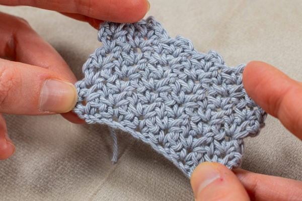 Gros plan de deux mains tenant un petit morceau de tissu crocheté gris clair avec un motif de point de ramage au crochet texturé, sur un fond neutre.