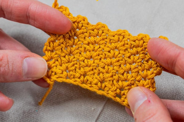Gros plan de deux mains tenant un petit morceau de tissu crocheté orange, présentant un motif texturé et tissé en point ramage au crochet, sur un fond de tissu gris clair.