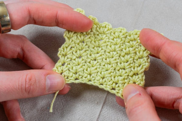 Gros plan des mains tenant un petit morceau de tissu crocheté jaune clair présentant le motif de point de ramage au crochet, sur un fond de tissu neutre.