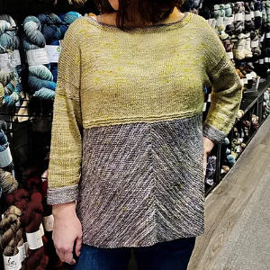 Une personne portant un pull tricoté bicolore à manches longues - jaune sur le dessus, gris sur le dessous - se tient devant des étagères remplies d'écheveaux de fils colorés dans un magasin de fils.