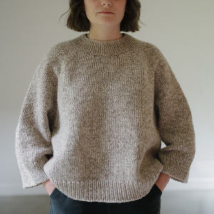 Une personne aux cheveux bruns mi-longs, vêtue d'un pull ample en maille beige et d'un pantalon sombre, se tient debout, les mains dans les poches, sur un fond clair uni.