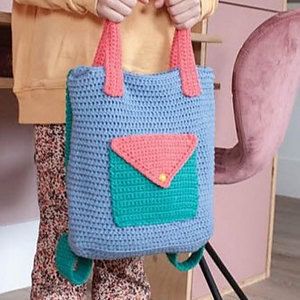Une personne tient un sac à dos crocheté avec un corps bleu, une poche avant verte, un rabat rose et des poignées roses. La personne porte un pantalon à motifs et un haut jaune ; une chaise et un bureau roses se trouvent à l'arrière-plan.