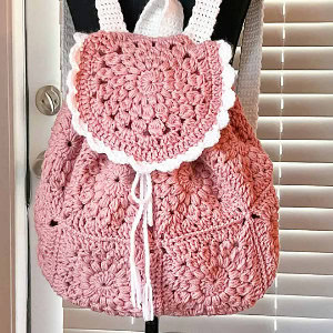 Un sac à dos crocheté rose avec un motif floral, un rabat festonné blanc et des cordons de serrage blancs est suspendu à un mannequin noir devant des stores et une porte.