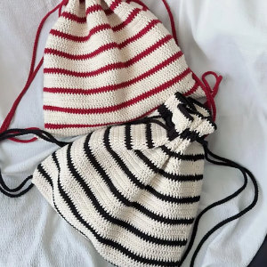 Deux sacs à cordon tricotés sur une surface blanche : l'un avec des rayures crème et rouges et un cordon rouge, l'autre avec des rayures crème et noires et un cordon noir.