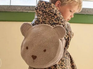 Un jeune enfant vêtu d'un manteau à motifs léopard porte un sac à dos circulaire en crochet en forme de visage d'ours, avec des yeux et un nez simples, debout à l'intérieur près d'une fenêtre.