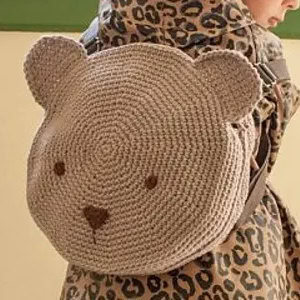Un enfant portant un sac à dos crocheté en forme d'ours, avec des oreilles rondes et un visage simple, par-dessus un manteau imprimé léopard.