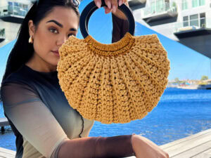 Une femme tient un sac à main rond, jaune, au crochet avec des poignées noires, debout en plein air au bord de l'eau, avec des immeubles modernes en arrière-plan.