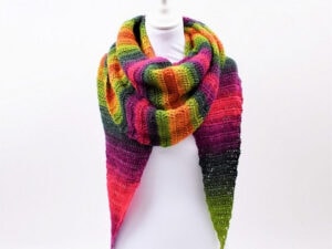 Un mannequin est drapé d'un grand châle triangulaire au crochet avec des rayures arc-en-ciel vibrantes en vert, jaune, orange, rouge, violet et rose - une inspiration parfaite si vous cherchez un tuto de châle au crochet facile. Le tout sur un fond blanc uni.