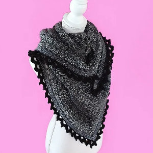 Un châle triangulaire gris avec une bordure festonnée noire est présenté sur un mannequin blanc sur un fond rose, une inspiration parfaite pour ceux qui cherchent un tuto de châle au crochet facile.
