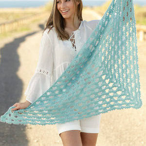 Une femme souriante, vêtue d'un chemisier blanc et d'un short, se tient sur une route ensoleillée et tend un grand châle bleu clair en dentelle au crochet - une inspiration parfaite pour tous ceux qui cherchent un tuto de châle au crochet facile.