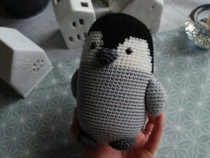 Une main tient un pingouin crocheté au corps gris, à la tête noire et au visage blanc. En arrière-plan, des maisons décoratives en céramique blanche et un plateau de table à motifs - une inspiration parfaite pour un tuto de pingouin au crochet en français.