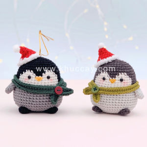 Deux pingouins crochetés portant des bonnets et des écharpes de Noël - l'un en rouge et gris, l'autre en vert et gris - se tiennent côte à côte sur un fond doucement éclairé par de faibles guirlandes lumineuses.