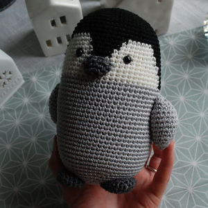 Une main tient un mignon pingouin crocheté au corps gris, au visage blanc, au bec noir et aux nageoires, sur fond de tissu à motifs et de maisons décoratives en céramique blanche.