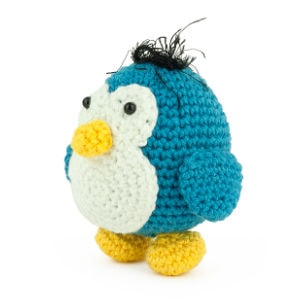 Un petit pingouin en peluche bleu et blanc, crocheté, avec des pieds jaunes, un bec jaune et une touffe de fil noir sur la tête, qui se tient debout sur un fond blanc.
