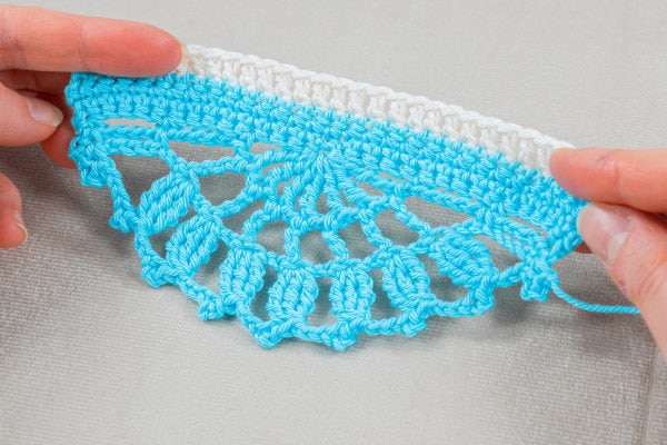 Deux mains tiennent un ouvrage au crochet représentant un motif semi-circulaire bleu et blanc avec des points ouverts en dentelle et une délicate bordure coquille au crochet sur un fond de tissu clair.