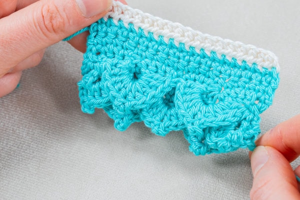 Gros plan de mains tenant une pièce crochetée avec une rangée supérieure blanche et un corps turquoise présentant un motif de coquille au crochet texturé, à bordure festonnée. L'arrière-plan est un tissu gris clair.
